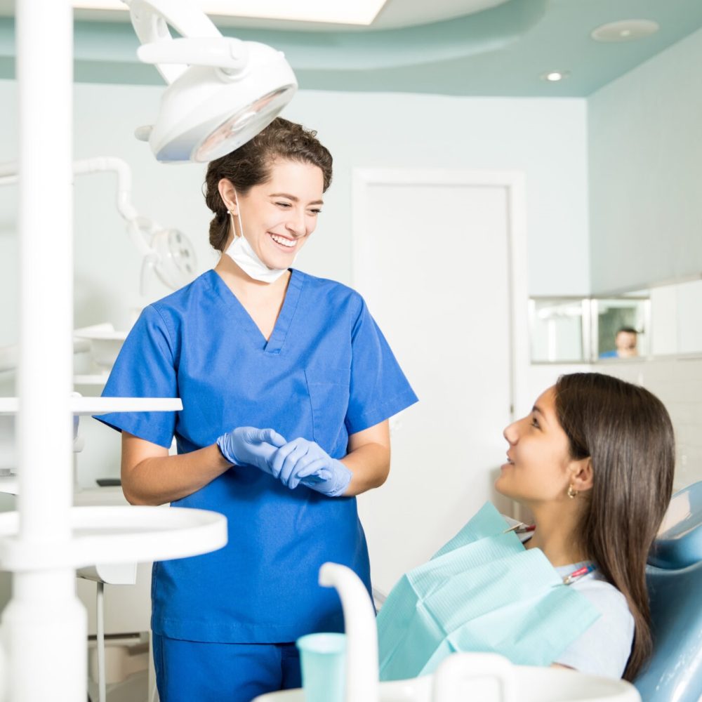 Smiling female dentist in uniform talking to teenage girl at dental clinic